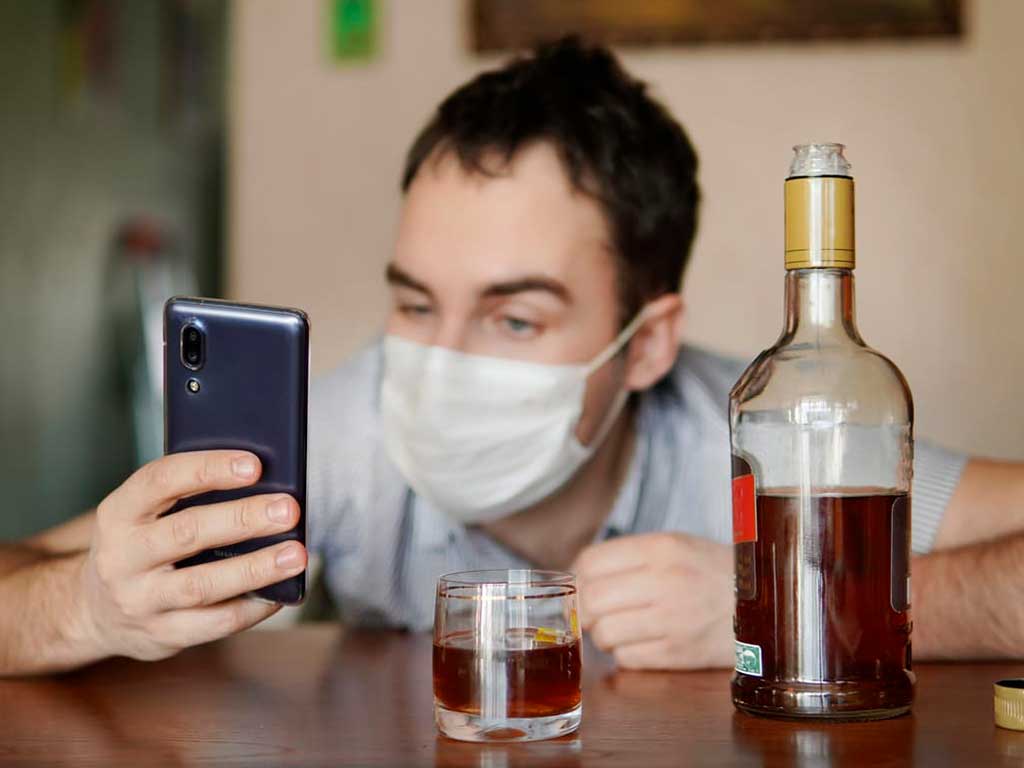 A man looking at his phone while a glass and bottle of alcohol are on the table