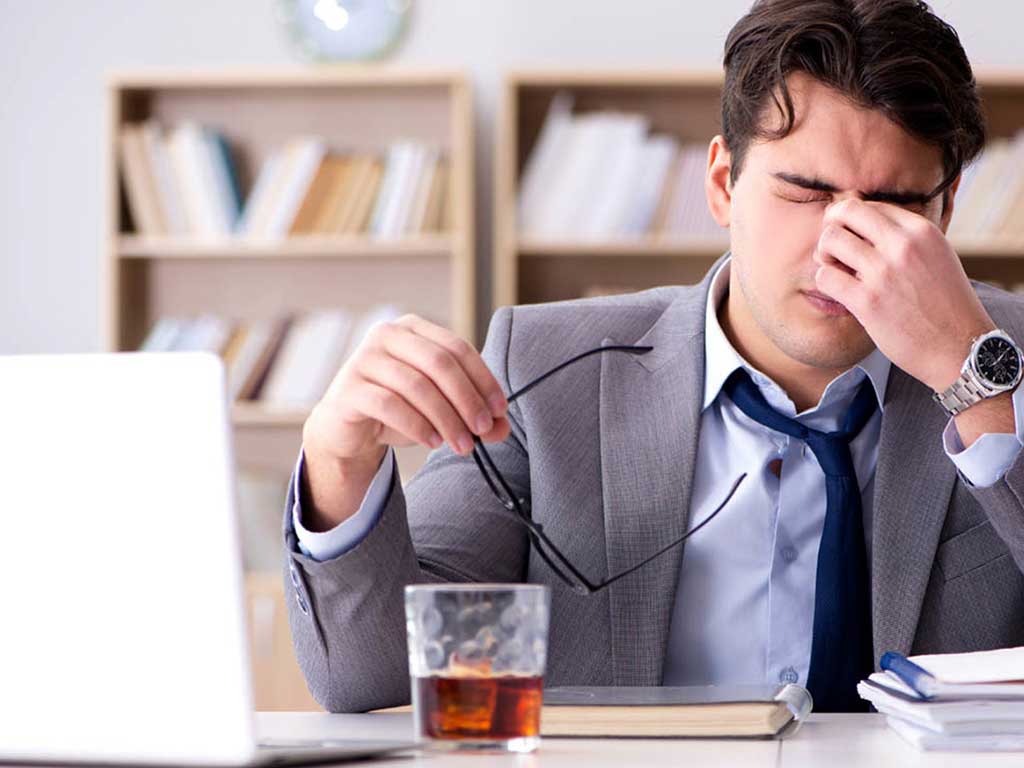A man grimacing while holding the bridge of his nose in front of his laptop and alcoholic drink