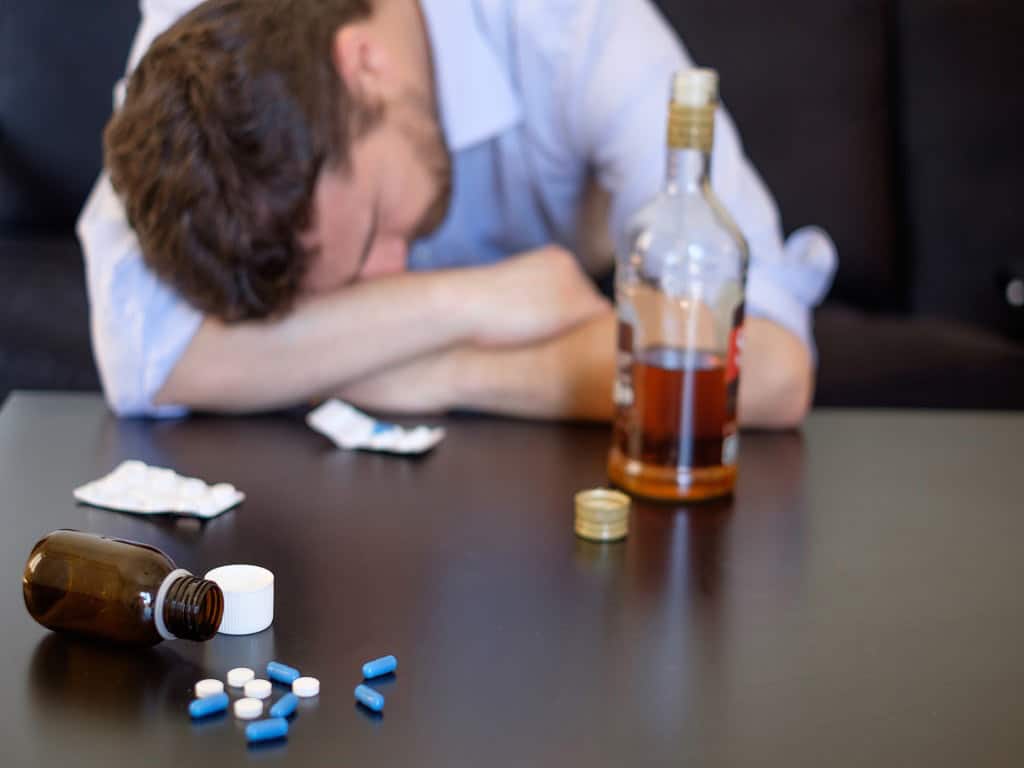 A man resting his head on a table while there are drugs and alcohol near him