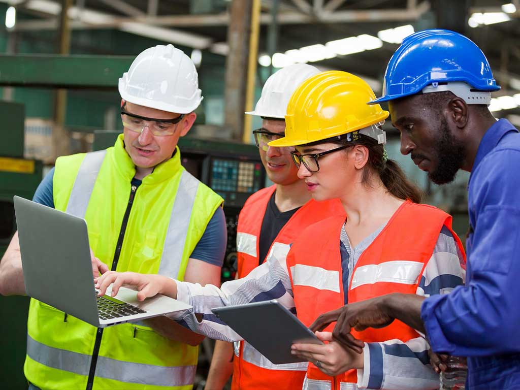 Four employees working while wearing hard hats and holding a tablet and a laptop