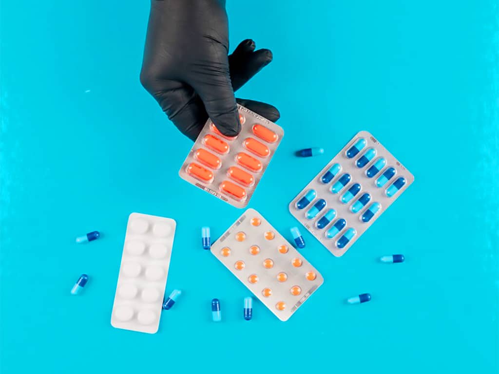 A hand holding a pack of pills while various types of drugs are scattered 
