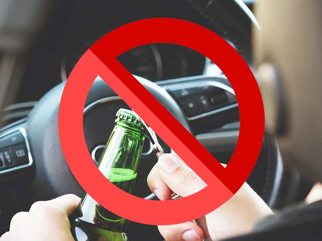A person behind the steering wheel opening a bottle of liquor with a red prohibition sign