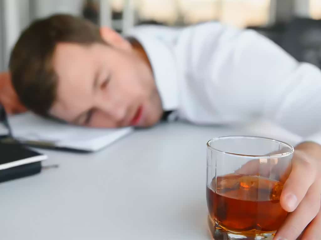 An employee sleeping on an office desk while holding a glass of alcohol